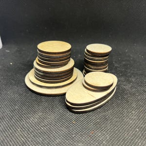 May include: Wooden craft supplies, including stacks of small, round, and oval wooden pieces. The pieces are light brown with darker brown edges, and are arranged in stacks of varying heights. The background is a dark gray.
