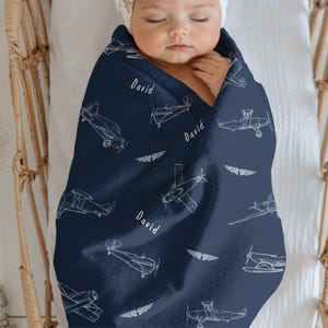 A navy blue Aviation Fleece Blanket for baby with a white airplane pattern and the name "David" printed on it. The blanket is wrapped around a baby in a crib. The blanket also features white wing designs.