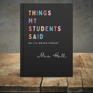 A black teacher journal gift featuring the text "THINGS MY STUDENTS SAID" in various colours. Underneath is the phrase "SO I'LL NEVER FORGET" and the name "Mrs. Hall" in cursive. The journal rests on a wooden surface.