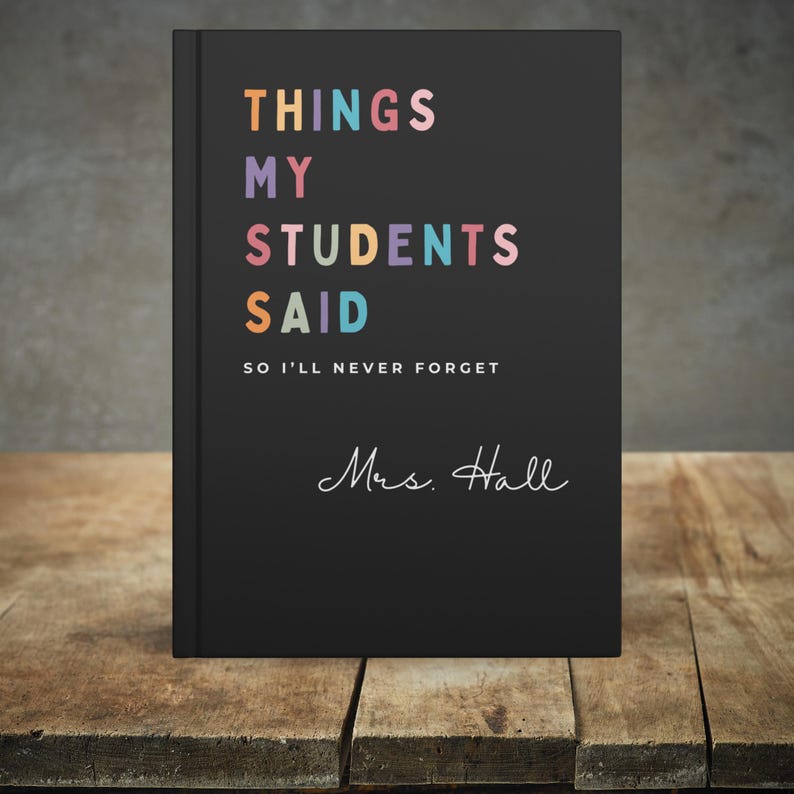 A black teacher journal gift featuring the text "THINGS MY STUDENTS SAID" in various colours. Underneath is the phrase "SO I'LL NEVER FORGET" and the name "Mrs. Hall" in cursive. The journal rests on a wooden surface.