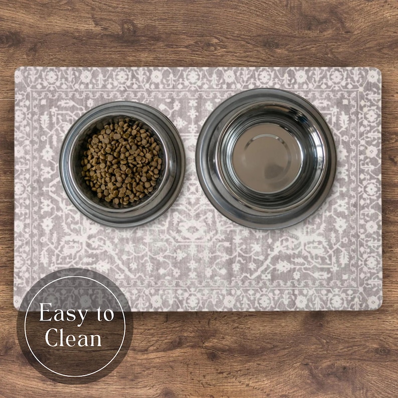 A pet feeding mat vintage boho style with two stainless steel bowls on a grey patterned mat. One bowl is filled with dry pet food, the other is empty. The mat has a decorative, vintage-style design.