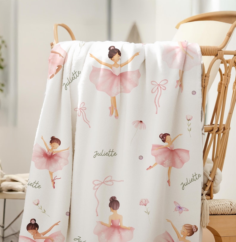 Kids Ballet Theme blanket with a pattern of pink-clad ballerinas, bows, and floral motifs. The name "Juliette" is written in cursive. The blanket is draped over a wicker chair, creating a soft, feminine aesthetic.
