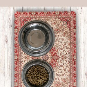 May include: An antique rug pet feeding station with two stainless steel bowls. One bowl is empty, and the other is filled with pet food. The rug has a red border and a cream-colored center with floral patterns. The text "Antique Rug PET FEEDING STATION" is at the top.