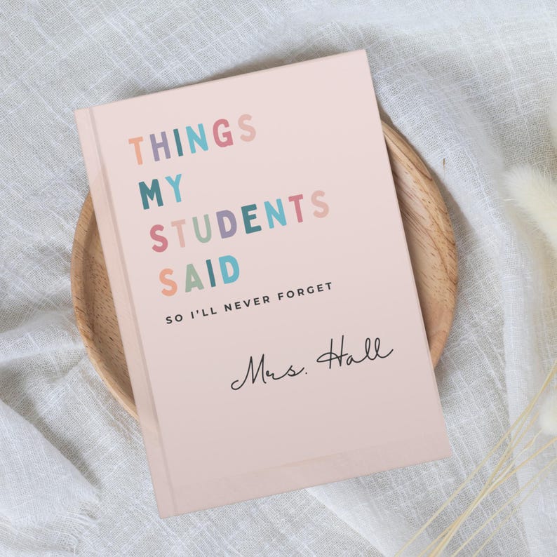 A pink gift for new teacher notebook with the text "THINGS MY STUDENTS SAID SO I'LL NEVER FORGET" in colourful letters. The notebook is personalised with the name "Mrs. Hall" in cursive. The notebook is on a wooden plate.