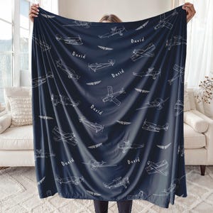 A navy blue  blanket Airplane Toddler Gift  with a repeating pattern of white airplane illustrations and the name "David". The blanket has a soft texture and is displayed against a neutral background. The design includes various airplane styles and wing motifs.