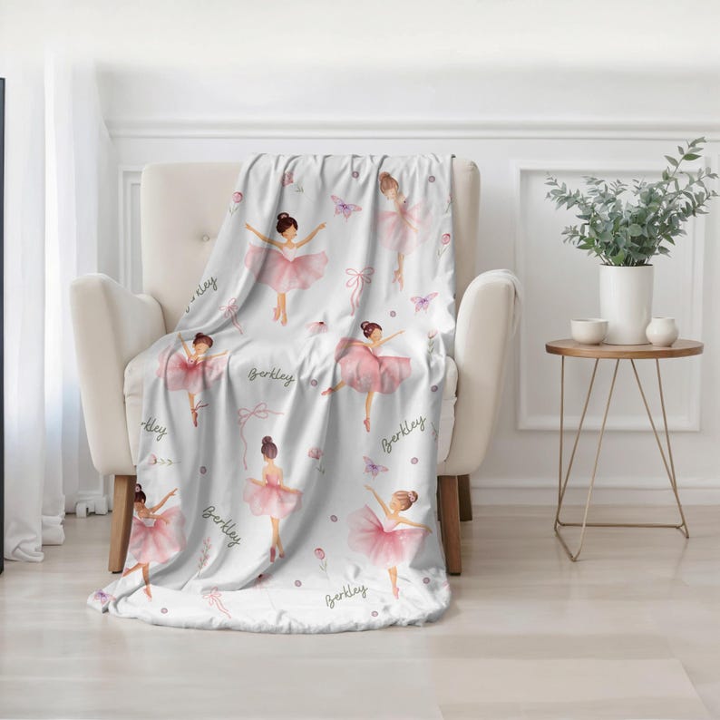 A white and pink personalized Ballet Theme Blanket is draped over a white chair in a contemporary-style room.