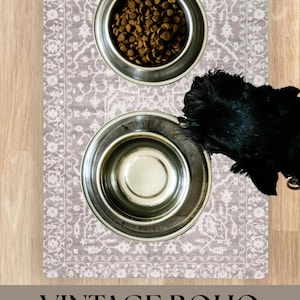 May include: A vintage boho pet feeding mat with two stainless steel bowls. One bowl is filled with dry pet food. The mat has a gray and white patterned design. A black dog is partially visible.