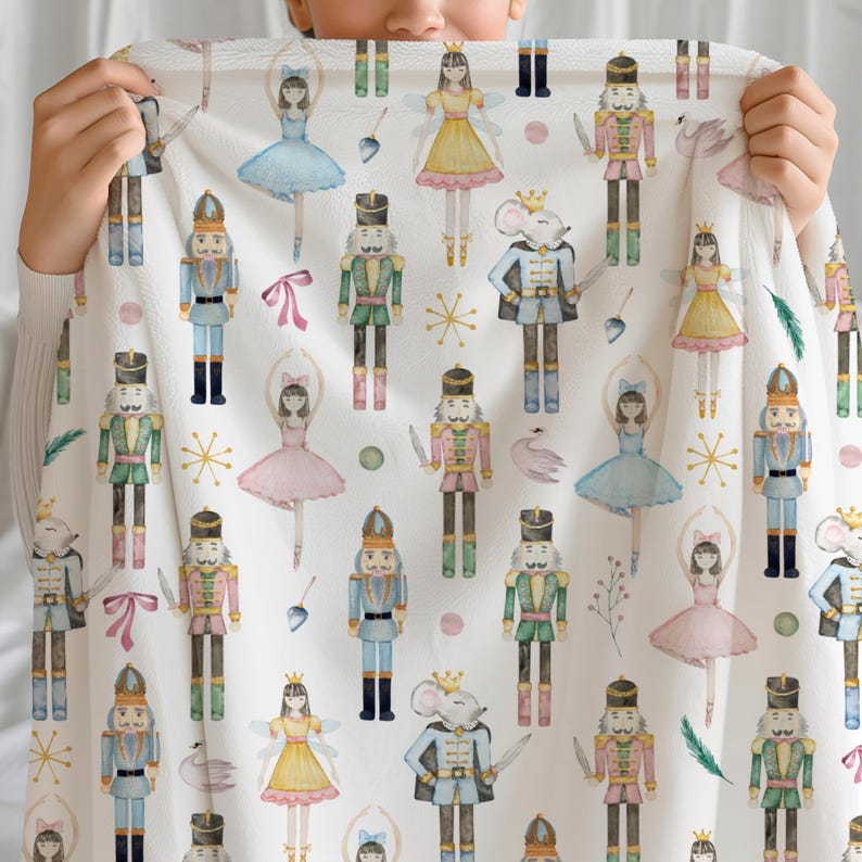 A nutcracker blanket ballet lover gift with a repeating pattern of watercolor-style nutcrackers and ballerinas in various colours. The blanket is held up, showing the full design. The design includes soldiers in green, blue, and gold uniforms, and ballerinas in pink, blue, and yellow dresses.