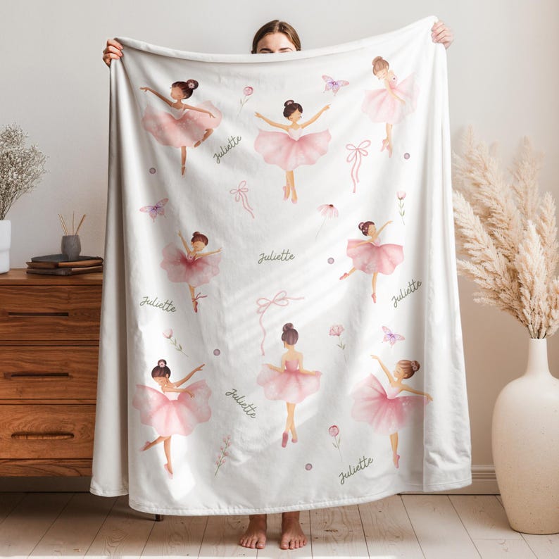 White blanket with a pattern of pink-clad ballerinas, bows, and butterflies. The name "Juliette" is written in cursive script. The blanket is held up by a person.