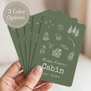 May include: A stack of rectangular olive green cards featuring white line art icons of a campfire, cabin, deer, pine trees, fishing rod, fish, mug, and lantern. The text reads Rush Family Cabin EST. 2024. A circular badge states 3 Color Options.