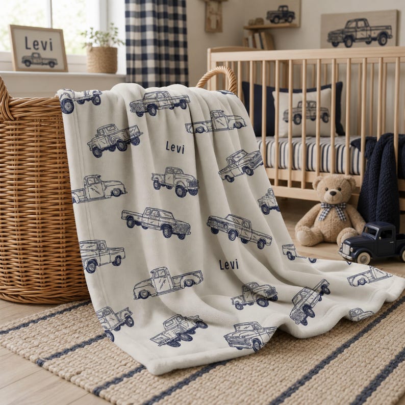A cream-coloured Baby Blanket for Boy Custom Name with a repeating pattern of blue vintage pickup trucks. The name "Levi" is printed in blue on the blanket. The blanket is draped over a wicker basket in a blue truck themed nursery