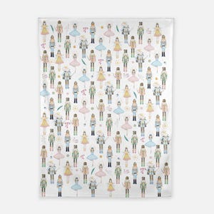 May include: A white fabric blanket featuring a repeating pattern of watercolor-style illustrations of nutcracker soldiers, ballerinas in pink, blue, and yellow dresses, and mouse characters, interspersed with small pink bows and gold star accents.