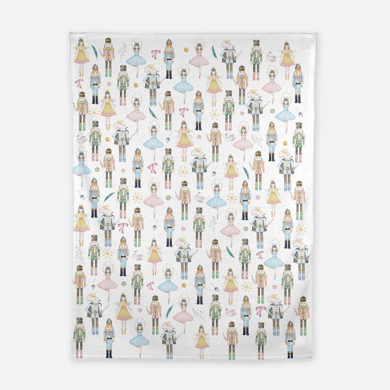 May include: A white fabric blanket featuring a repeating pattern of watercolor-style illustrations of nutcracker soldiers, ballerinas in pink, blue, and yellow dresses, and mouse characters, interspersed with small pink bows and gold star accents.