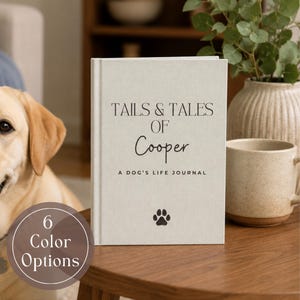 May include: A light beige hardback journal with the text TAILS & TALES OF Cooper A DOG'S LIFE JOURNAL printed in black, featuring a small black paw print icon at the bottom centre. The journal is positioned on a wooden surface next to a mug.