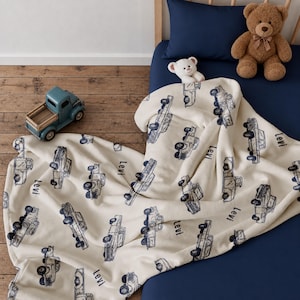 A Cream-coloured Vintage Truck Toddler Gift blanket featuring a repeating pattern of blue trucks and the name "Levi". The blanket is draped over a toddler bed. A blue truck is  on the wooden floor.