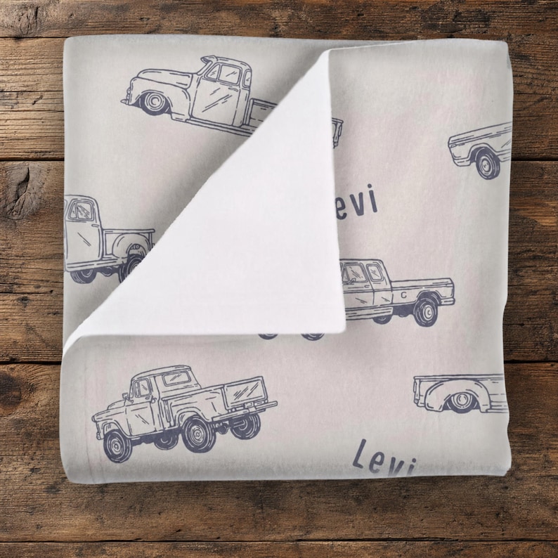 K&ouml;nnte beinhalten: Eine hellgraue Decke mit einem sich wiederholenden Muster aus dunkelblauen Strichzeichnungen von Oldtimer-Pickups. Der Stoff ist umgeschlagen und zeigt eine einfarbig wei&szlig;e Unterseite. Der Name Levi ist in dunkelblauer Schrift aufgedruckt.