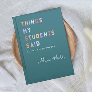 A teal teacher Christmas Gift journal with the text "THINGS MY STUDENTS SAID" in colourful letters. Below the title, it reads "SO I'LL NEVER FORGET" and "Mrs. Hall" in cursive. The journal is on a wooden plate.