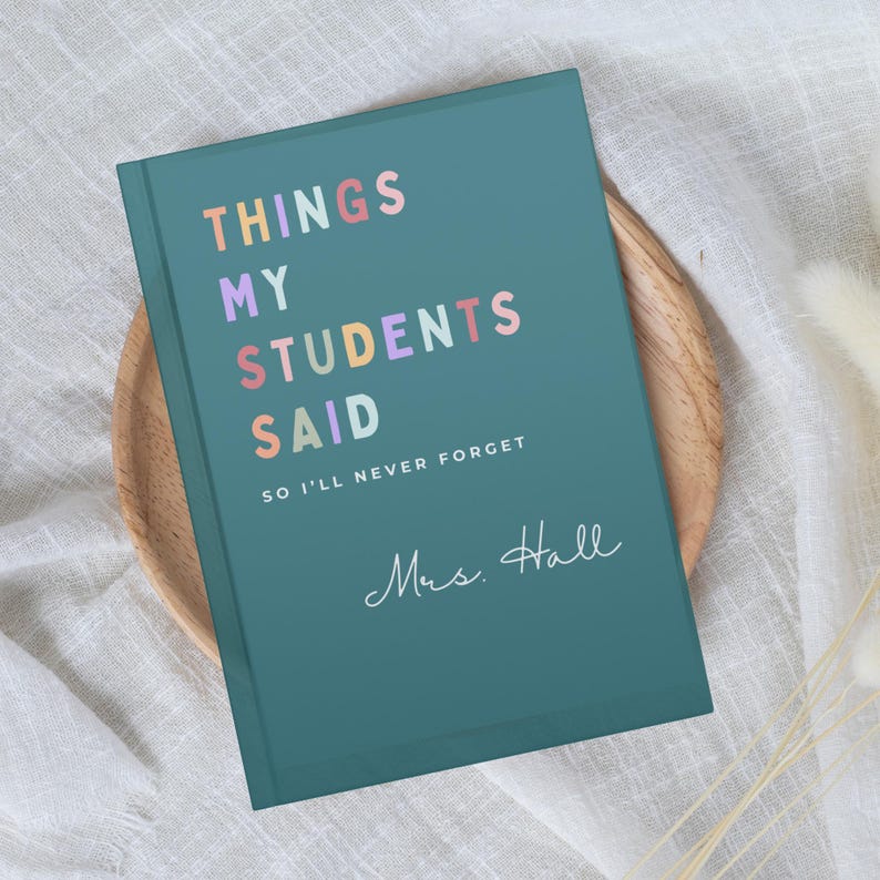 A teal teacher Christmas Gift journal with the text "THINGS MY STUDENTS SAID" in colourful letters. Below the title, it reads "SO I'LL NEVER FORGET" and "Mrs. Hall" in cursive. The journal is on a wooden plate.