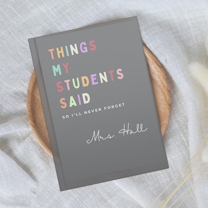 A grey teacher notebook with the text "THINGS MY STUDENTS SAID" in colourful letters. Below the title is the text "SO I'LL NEVER FORGET" and the name "Mrs. Hall" in cursive. The notebook is on a wooden plate.
