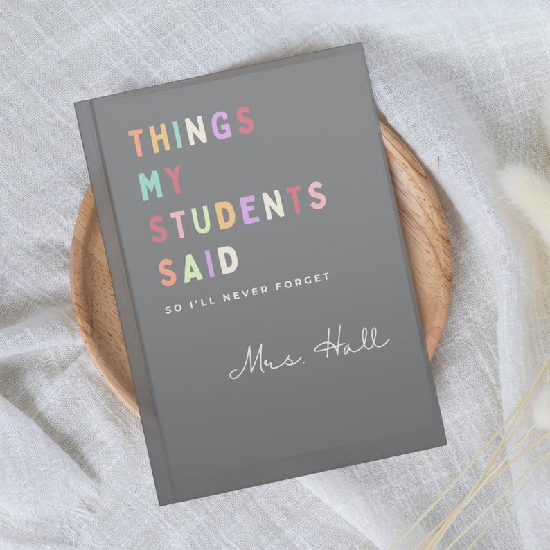A grey teacher notebook with the text "THINGS MY STUDENTS SAID" in colourful letters. Below the title is the text "SO I'LL NEVER FORGET" and the name "Mrs. Hall" in cursive. The notebook is on a wooden plate.