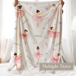 A woman is holding a Custom Ballerina Name Blanket for Girls featuring pink ballerinas, flowers, and butterflies. The blanket is customized with a name in script font.