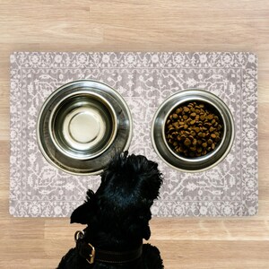A black dog with a brown collar gazes at two stainless steel dog bowls on a Pet Feeding Station Dog Bowl Rug. One bowl is empty, the other filled with brown kibble. The mat features a floral pattern.