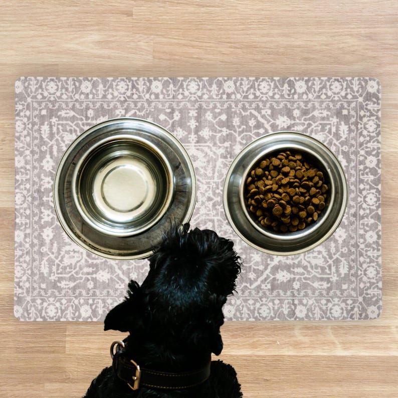 A black dog with a brown collar gazes at two stainless steel dog bowls on a Pet Feeding Station Dog Bowl Rug. One bowl is empty, the other filled with brown kibble. The mat features a floral pattern.