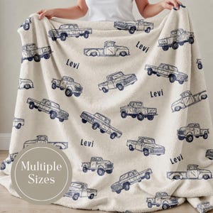 Cream-coloured Personalized truck blanket for boy with a repeating pattern of navy blue vintage pickup trucks. The name "Levi" is printed in navy blue across the blanket. The blanket is made of a soft, plush material.