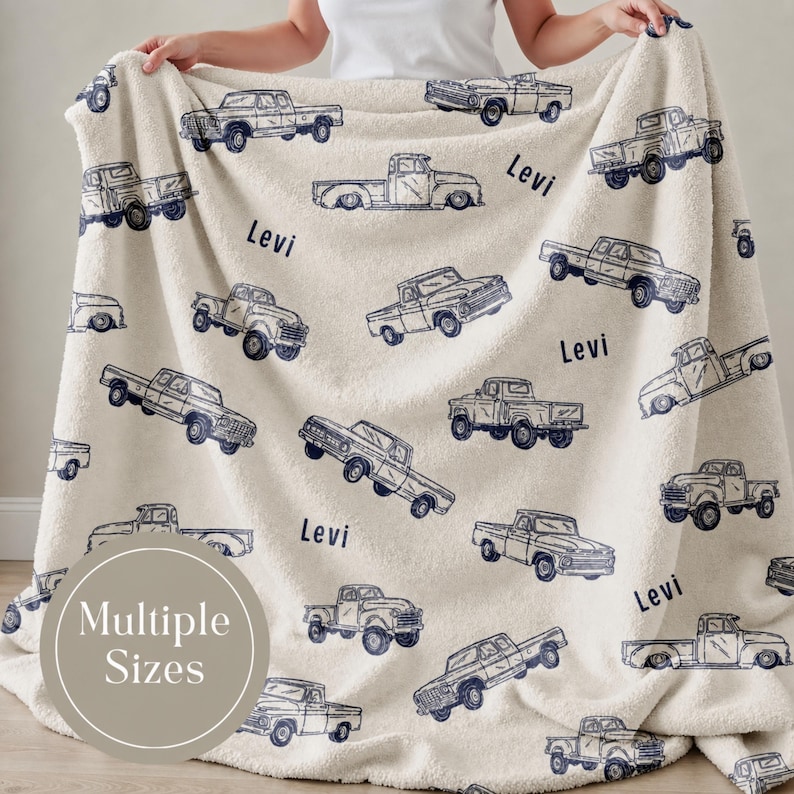 Cream-coloured Personalized truck blanket for boy with a repeating pattern of navy blue vintage pickup trucks. The name "Levi" is printed in navy blue across the blanket. The blanket is made of a soft, plush material.