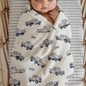 A cream-coloured Custom Name Baby Boy Blanket  featuring a navy blue pattern of vintage pickup trucks. The name "Levi" is printed on the blanket. A baby is wrapped in the blanket inside a bassinet.