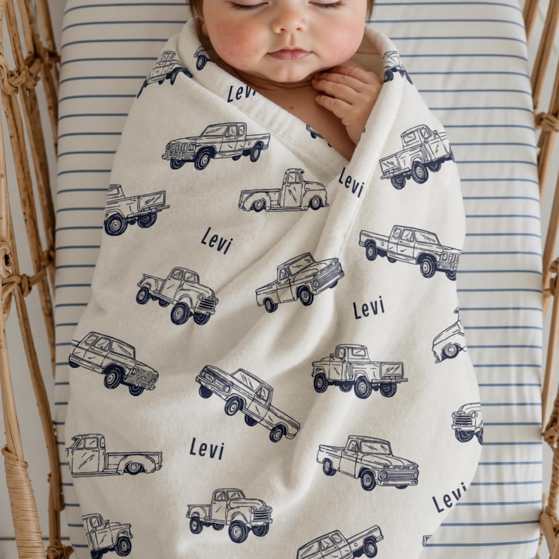 A cream-coloured Custom Name Baby Boy Blanket  featuring a navy blue pattern of vintage pickup trucks. The name "Levi" is printed on the blanket. A baby is wrapped in the blanket inside a bassinet.
