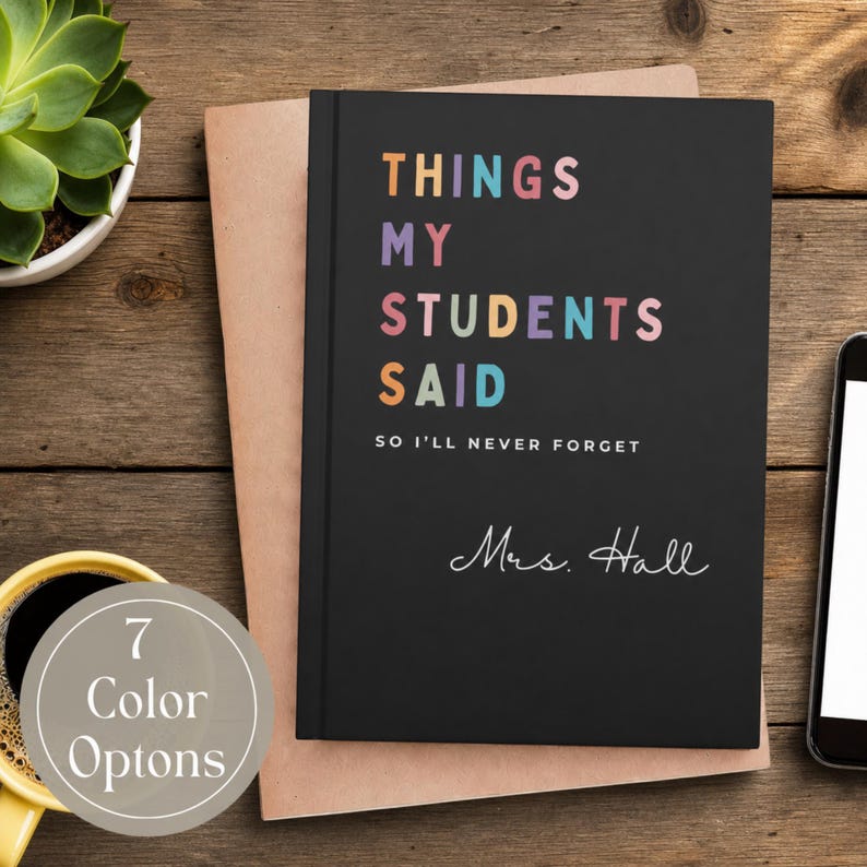 A personalized teacher journal keepsake with the text "THINGS MY STUDENTS SAID" in colourful letters. Below the text is "SO I'LL NEVER FORGET" and the name "Mrs. Hall" in cursive. The notebook is on a wooden surface with a cup of coffee and a smartphone.