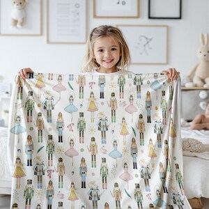 A Pink Christmas Cottagecore Throw for Her with a repeating pattern of watercolour-style nutcrackers, ballerinas, and mice. The blanket has a soft, cosy appearance, suitable for a child's bedroom. The design includes various colours such as blue, pink, yellow, and green.