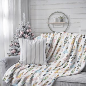 A nutcracker Christmas Decor throw blanket with a colourful Nutcracker and ballerina pattern is draped over a grey sofa. A striped pillow and a decorated Christmas tree are also visible. The blanket has a soft, plush texture.