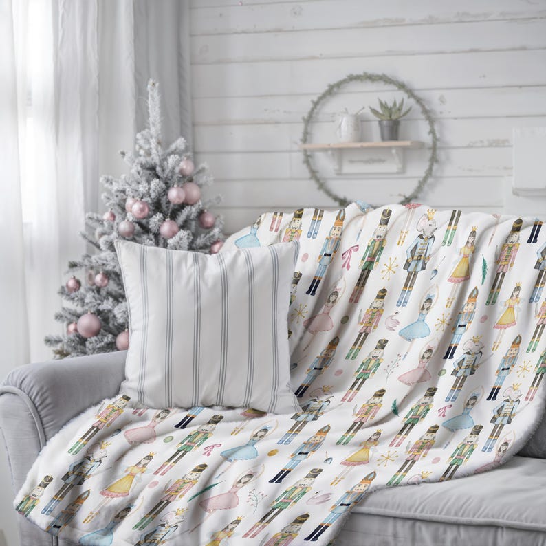 A nutcracker Christmas Decor throw blanket with a colourful Nutcracker and ballerina pattern is draped over a grey sofa. A striped pillow and a decorated Christmas tree are also visible. The blanket has a soft, plush texture.