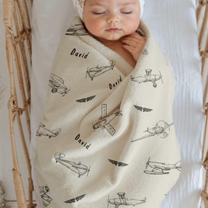 Cream Custom Name Vintage Airplane baby blanket featuring black airplane illustrations and the name "David". The blanket is wrapped around a baby in a crib. The blanket has various airplane designs and wing emblems.