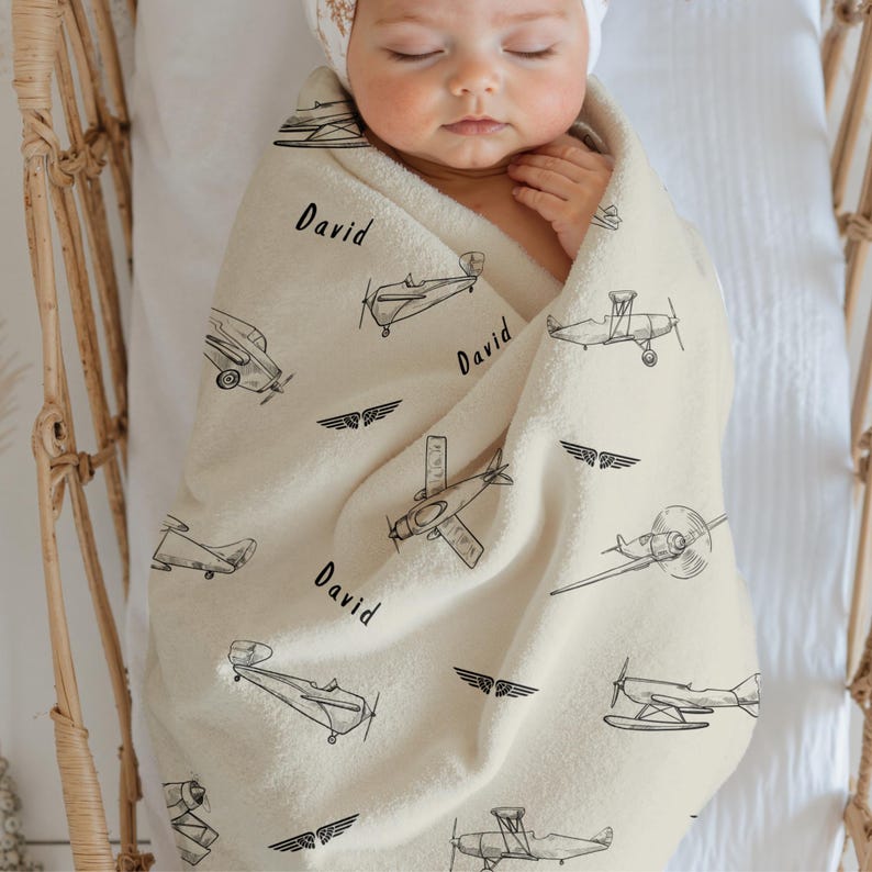 Cream Custom Name Vintage Airplane baby blanket featuring black airplane illustrations and the name "David". The blanket is wrapped around a baby in a crib. The blanket has various airplane designs and wing emblems.