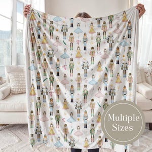 May include: A white blanket featuring a repeating pattern of watercolor-style nutcrackers and ballerinas in various colors. The blanket has a soft, inviting appearance, perfect for adding a touch of whimsy to any room. The blanket is available in multiple sizes.