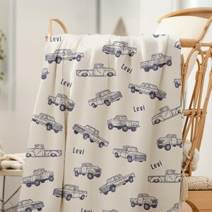 A cream-coloured Truck Theme Boy Gift Blanket with a repeating pattern of blue vintage pickup trucks. The name "Levi" is printed on the blanket. The blanket is draped over basket.
