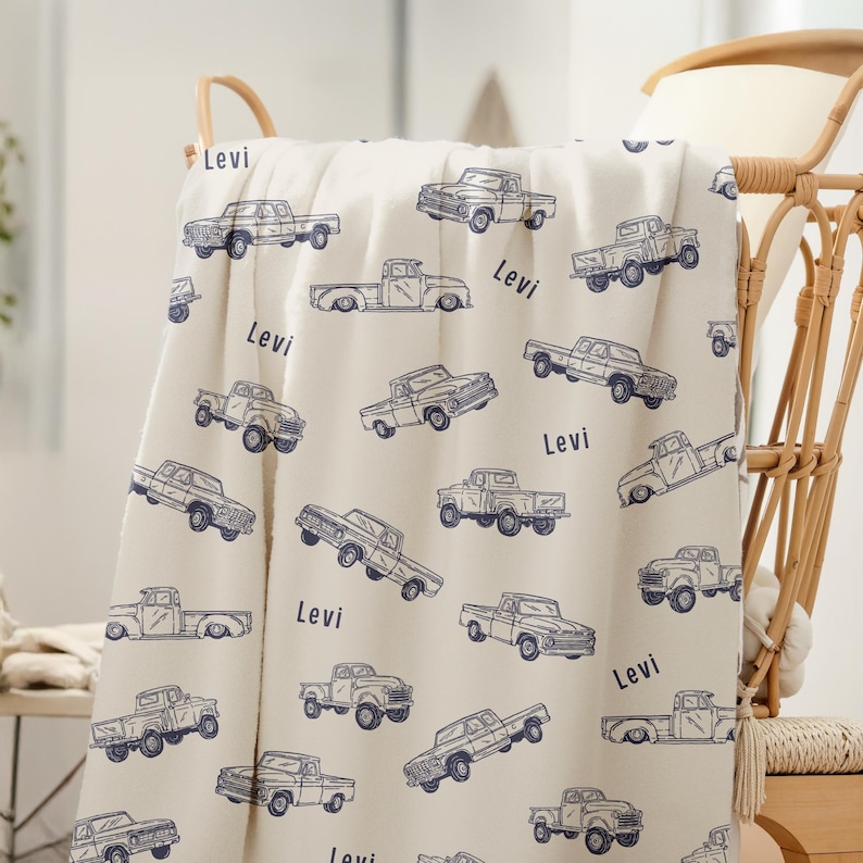 A cream-coloured Truck Theme Boy Gift Blanket with a repeating pattern of blue vintage pickup trucks. The name "Levi" is printed on the blanket. The blanket is draped over basket.