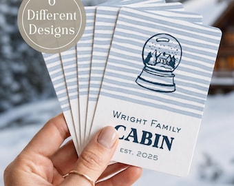 May include: A set of rectangular cards featuring a light blue and white horizontal striped pattern. The top card displays a dark blue snow globe illustration with a cabin inside, and the text WRIGHT FAMILY CABIN EST. 2025. A circular badge states 6 Different Designs.