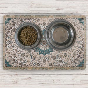 A pet feeding station with two stainless steel bowls on a patterned mat. One bowl contains dry pet food, the other is empty. The mat has a cream, teal, and brown design, with a detailed, ornate pattern.