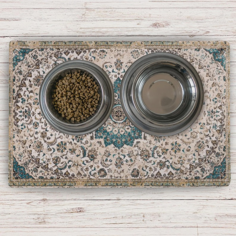 A pet feeding station with two stainless steel bowls on a patterned mat. One bowl contains dry pet food, the other is empty. The mat has a cream, teal, and brown design, with a detailed, ornate pattern.