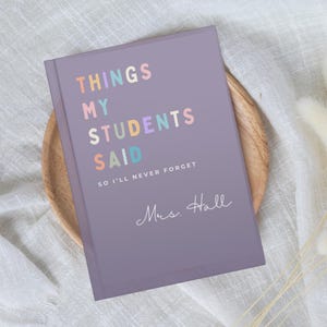 A lilac Teacher Appreciation Gift notebook with the text "THINGS MY STUDENTS SAID" in pastel colours. Below the title is the text "SO I'LL NEVER FORGET" and the name "Mrs. Hall" in cursive. The notebook is on a wooden plate.