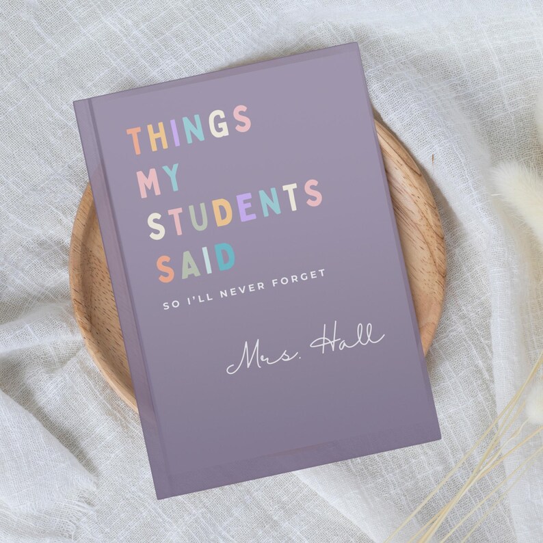 A lilac Teacher Appreciation Gift notebook with the text "THINGS MY STUDENTS SAID" in pastel colours. Below the title is the text "SO I'LL NEVER FORGET" and the name "Mrs. Hall" in cursive. The notebook is on a wooden plate.