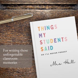 A white custom teacher gift notebook with the text "THINGS MY STUDENTS SAID" in colourful letters. The notebook also includes the text "SO I'LL NEVER FORGET" and a handwritten signature. A gold pen rests on a wooden surface.