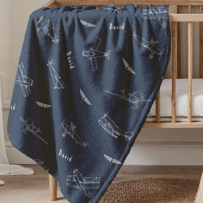 A navy blue Airplane Nursery Decor baby blanket featuring a white airplane and wing design. The name "David" is printed in white on the blanket. The blanket is draped over a wooden crib.