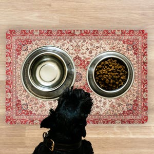 Pet Food Bowl Mat featuring a Persian rug pattern in red, cream, and beige. Two stainless steel dog bowls are on the mat, one empty and the other filled with brown kibble. A black dog with a brown collar is looking at the bowls.