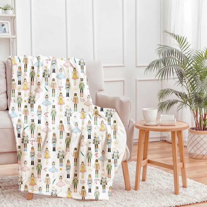 A nutcracker decor blanket with a repeating pattern of nutcrackers and ballerinas in various colours, draped over a beige armchair. The design includes figures in green, yellow, blue, and pink. The blanket is made of a soft material, suitable for a cosy home.