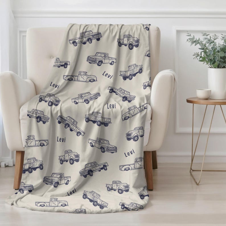 A beige Personalized Truck Blanket for Boy with a pattern of blue vintage pickup trucks and the name "Levi". The blanket is draped over a white armchair. A cozy home decor item, perfect for adding a touch of style to any room.
